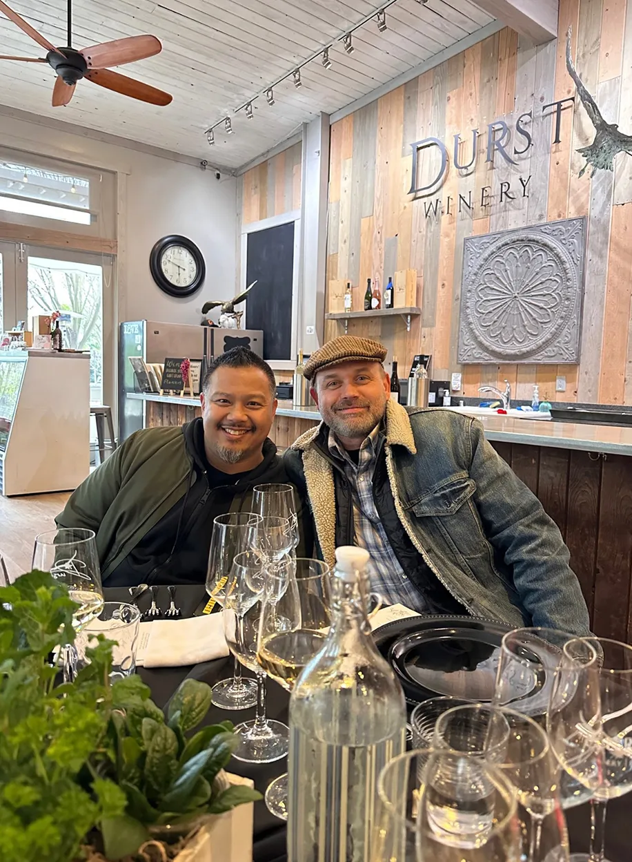 Two team members smiling together at a winery tasting bar with wine glasses on the table