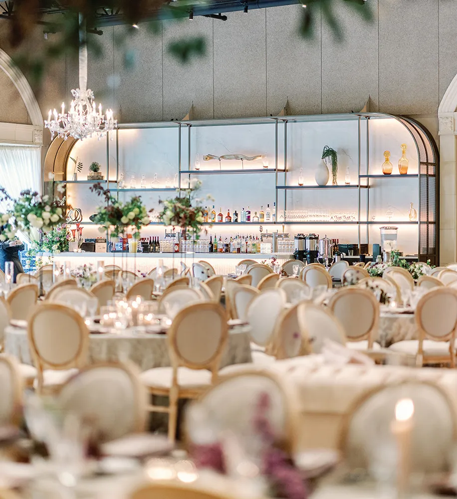Elegant indoor reception hall with round tables, gold-trimmed chairs, crystal chandeliers, and an arched bar with glass shelving