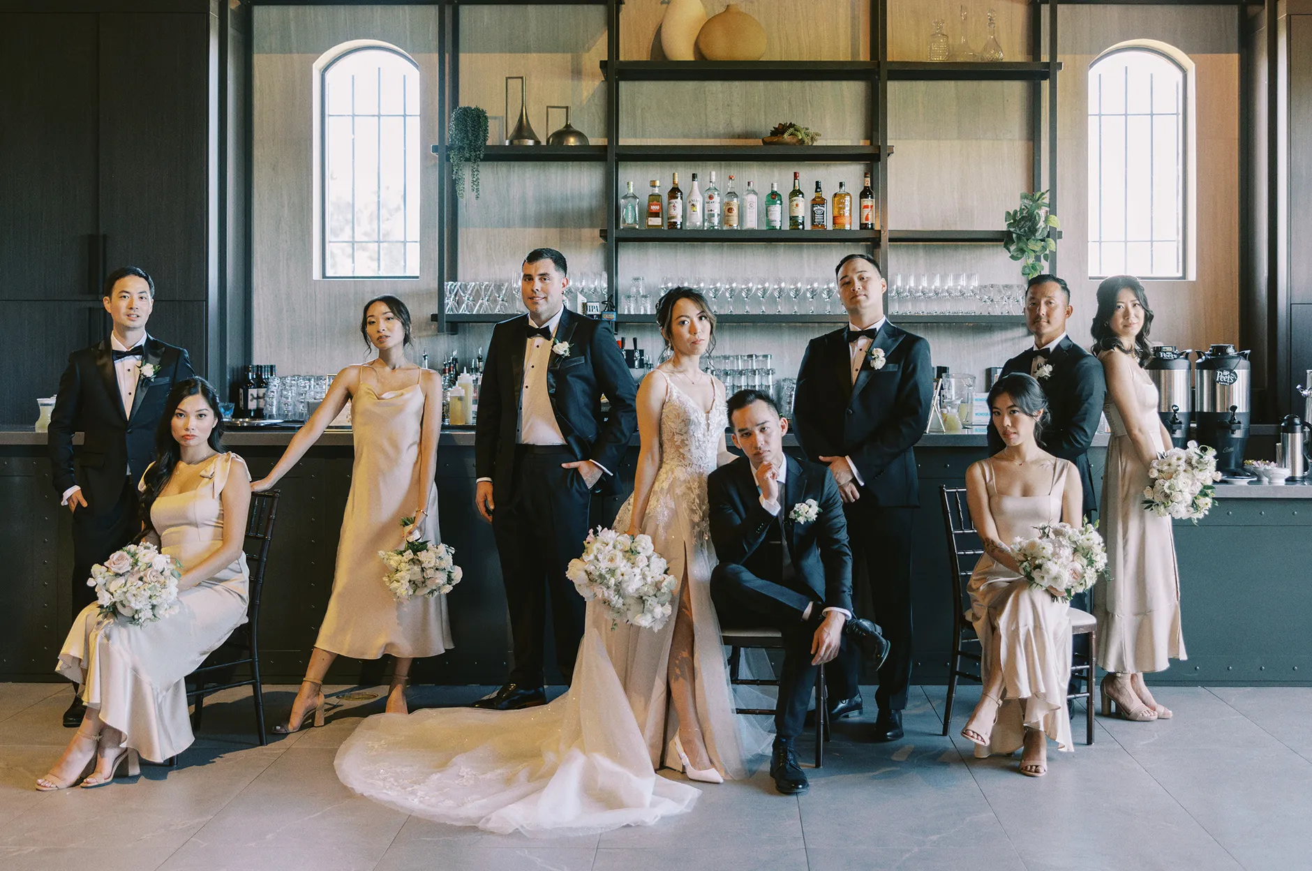 Wedding party in black tuxedos and champagne dresses posing together in front of the venue bar