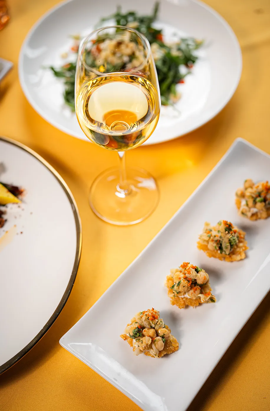 Glass of white wine surrounded by plated arugula salad and crispy herb-topped canapés on a golden tablecloth