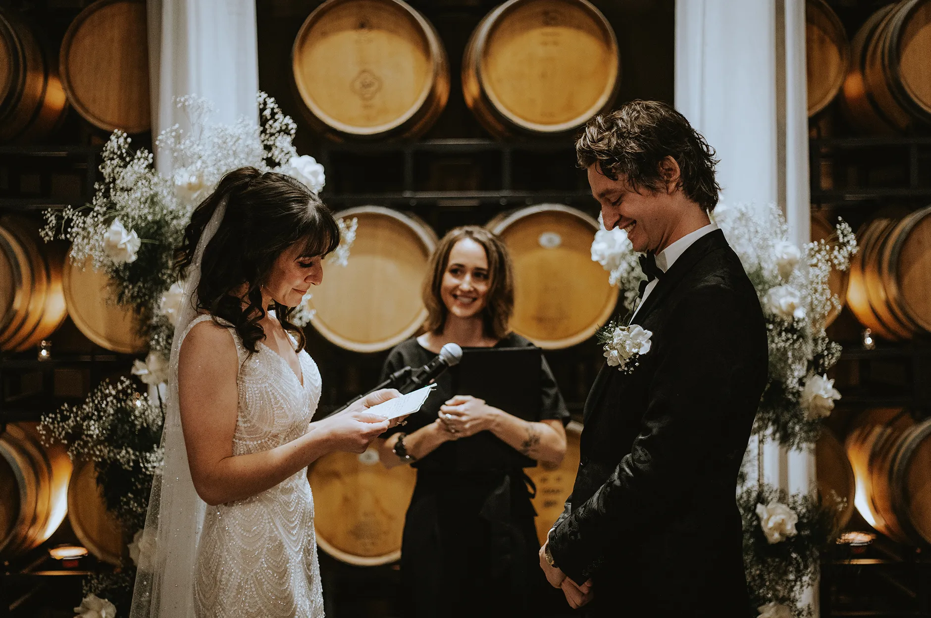 Bride reading her vows to the groom during a ceremony framed by wine barrels and white floral arrangements