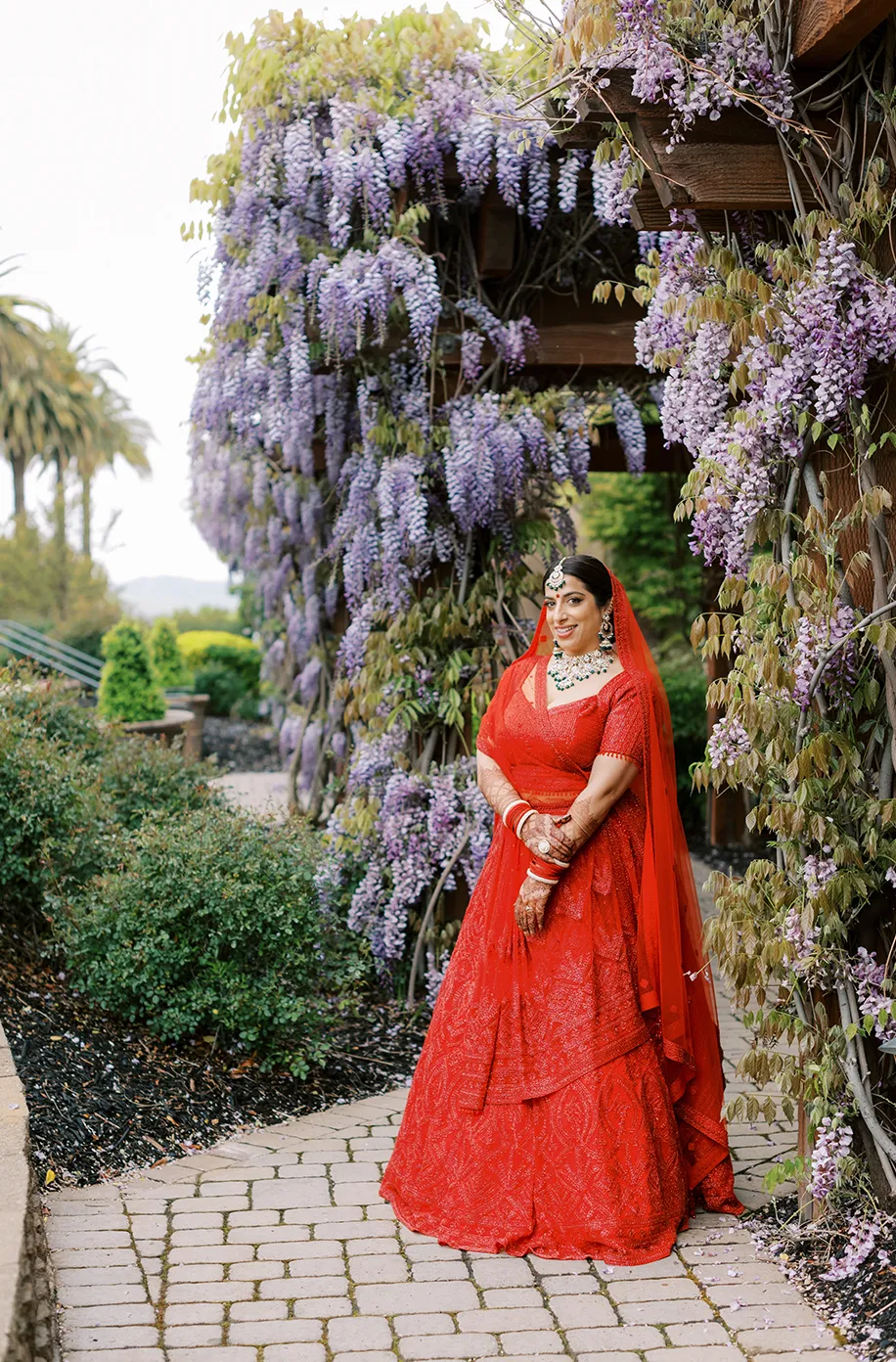 Bride in a red lehenga posing beneath a cascading wisteria arbor with palm trees in the background