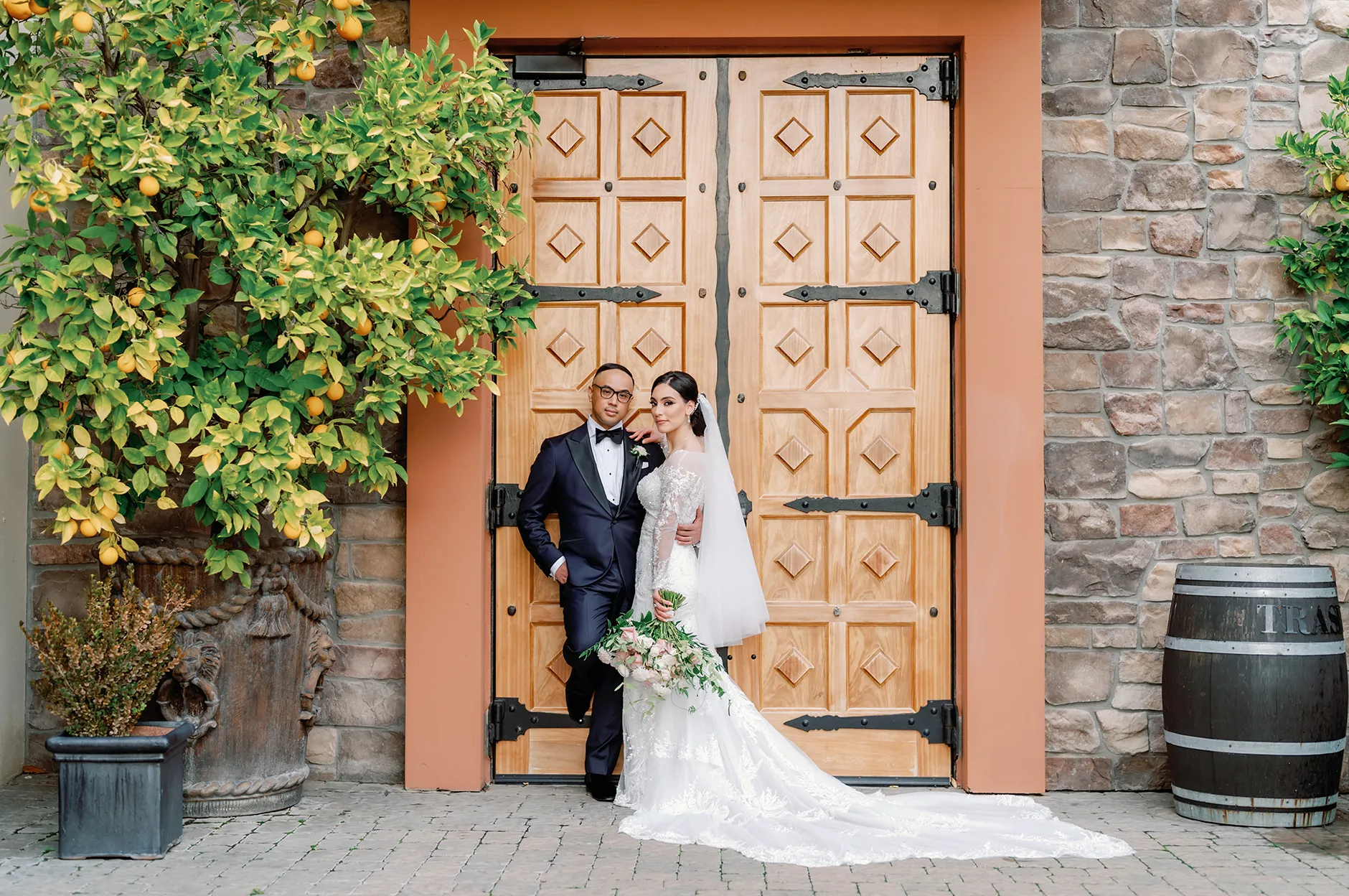 Bride and groom posing in front of large rustic wooden doors framed by stone walls, a citrus tree, and a wine barrel