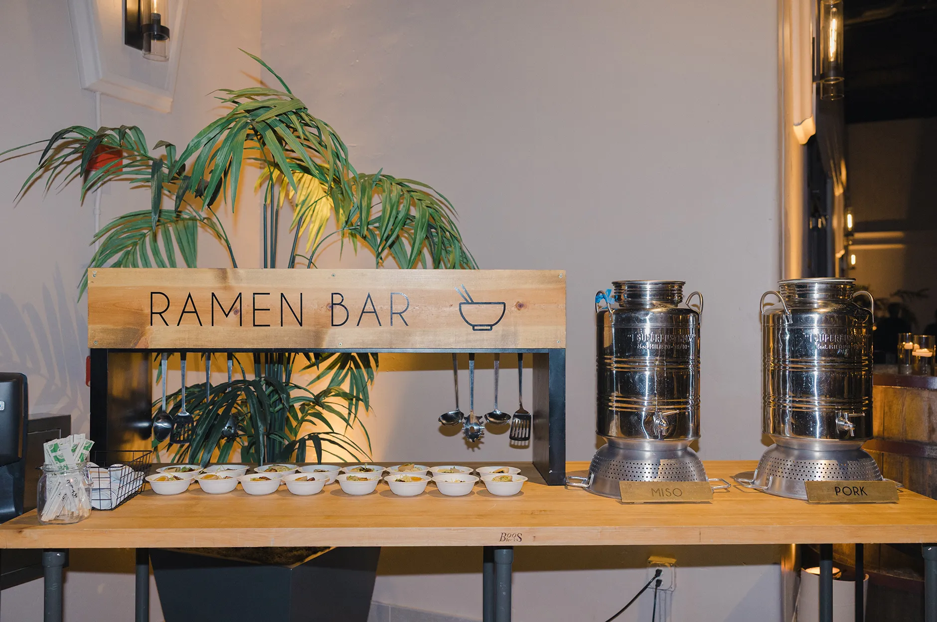Ramen bar station with a wooden sign, rows of small tasting bowls, and stainless steel broth dispensers
