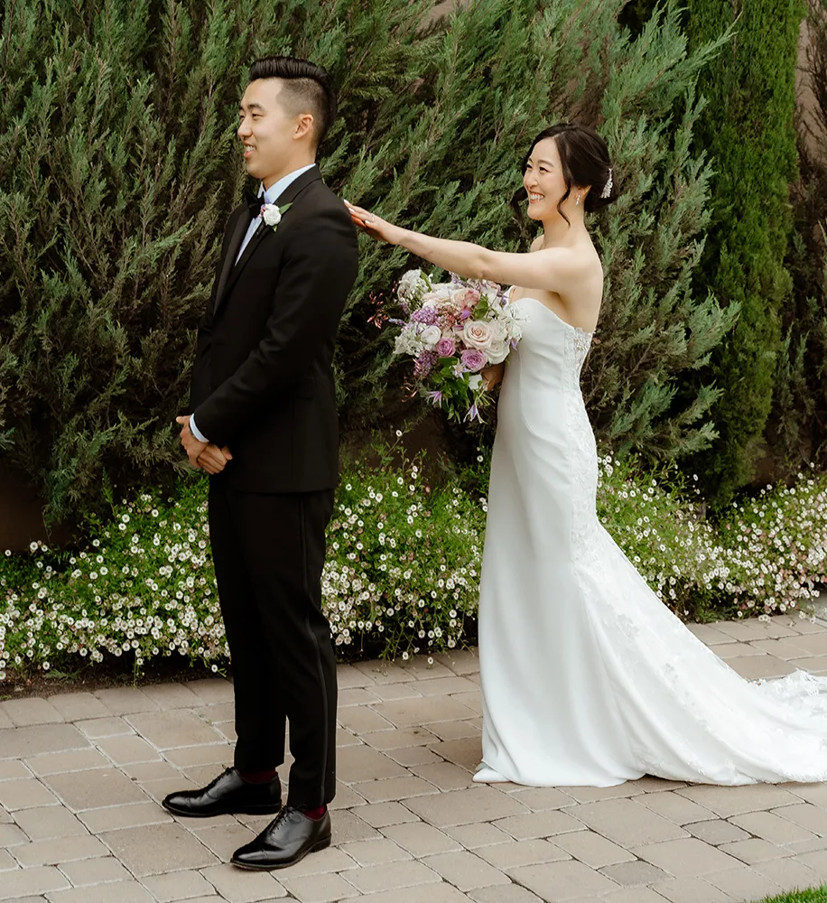 Bride tapping the groom on the shoulder for a first-look reveal in a garden with cypress trees and white flowers