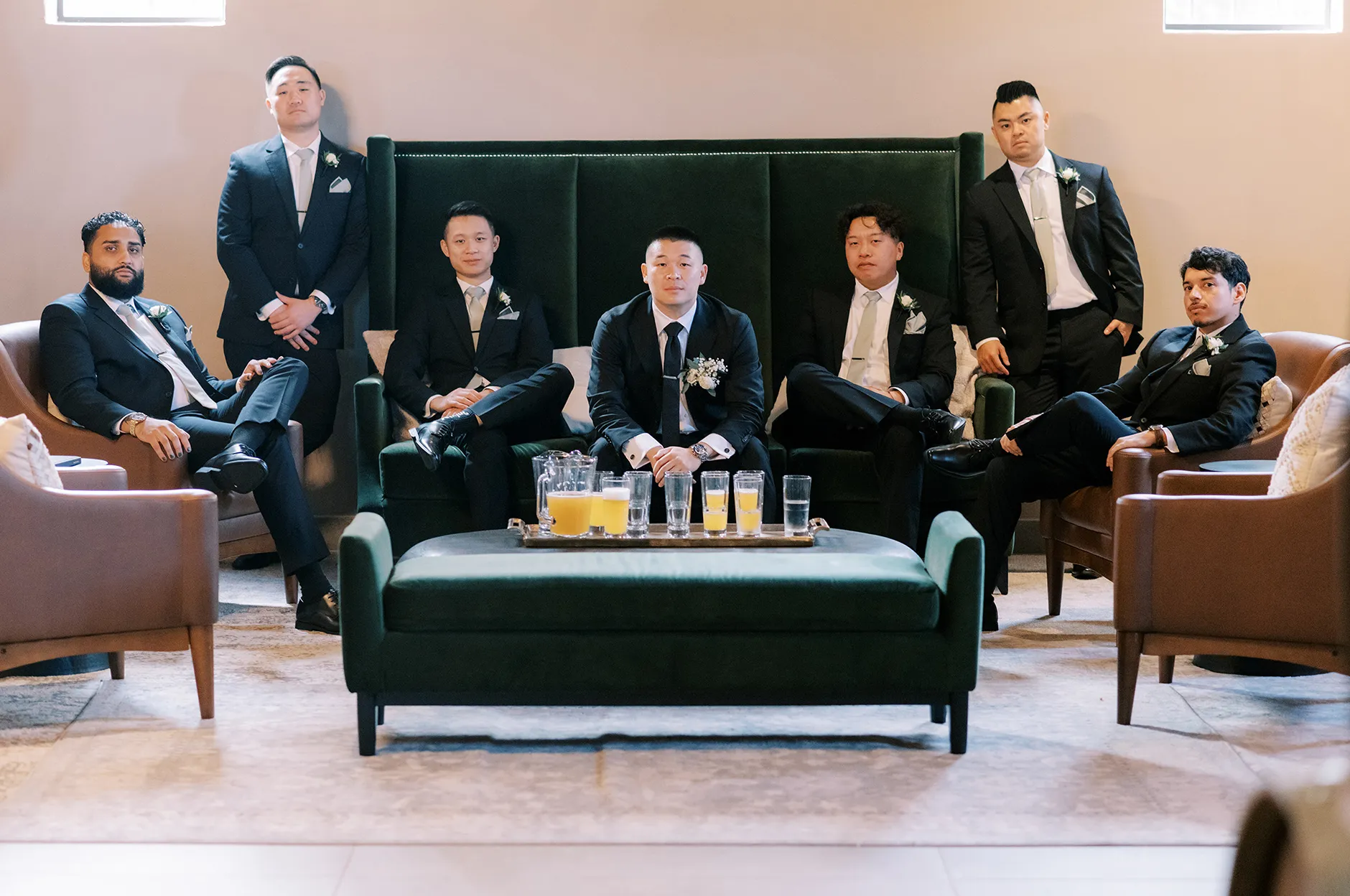 Groom and groomsmen in dark suits seated on a green velvet sofa in the venue lounge with cocktails on the table