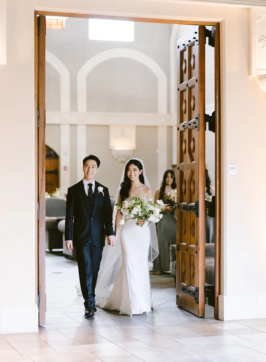 Bride and groom walking through ornate carved wooden doors into the sunlit venue interior with arched windows