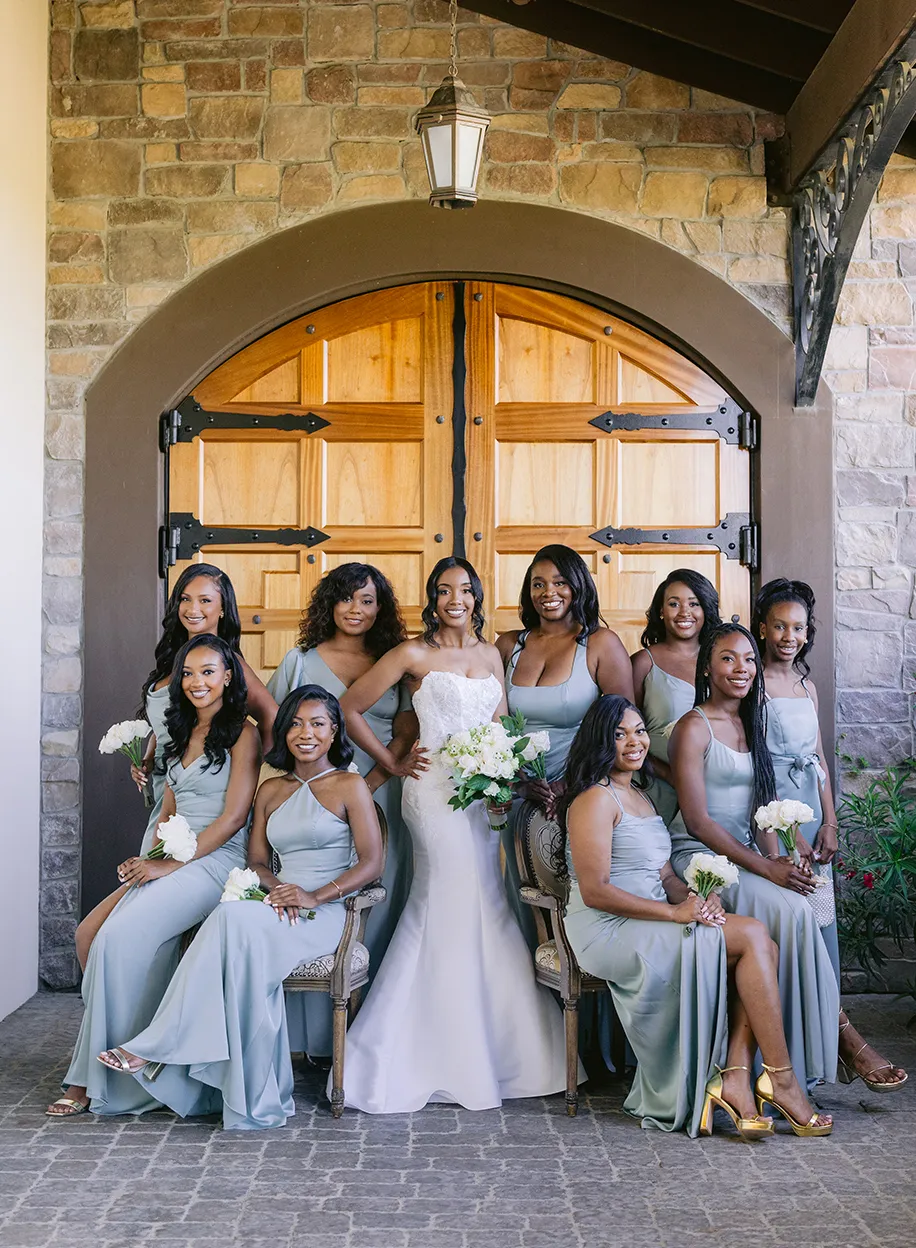 Bride and bridesmaids in dusty blue dresses posing together beneath a stone archway with wooden doors