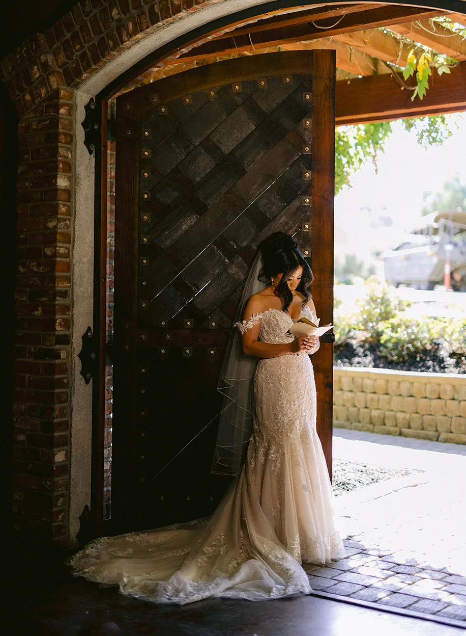 Bride reading a letter while standing in a large arched wooden doorway bathed in afternoon sunlight