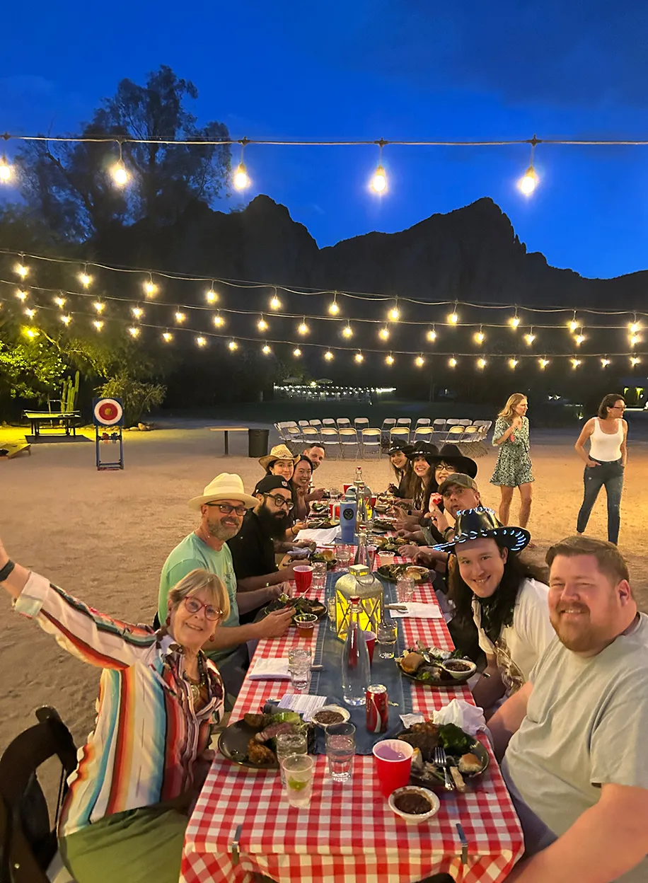 Team dinner at a long checkered-tablecloth table under string lights with desert cliffs at twilight