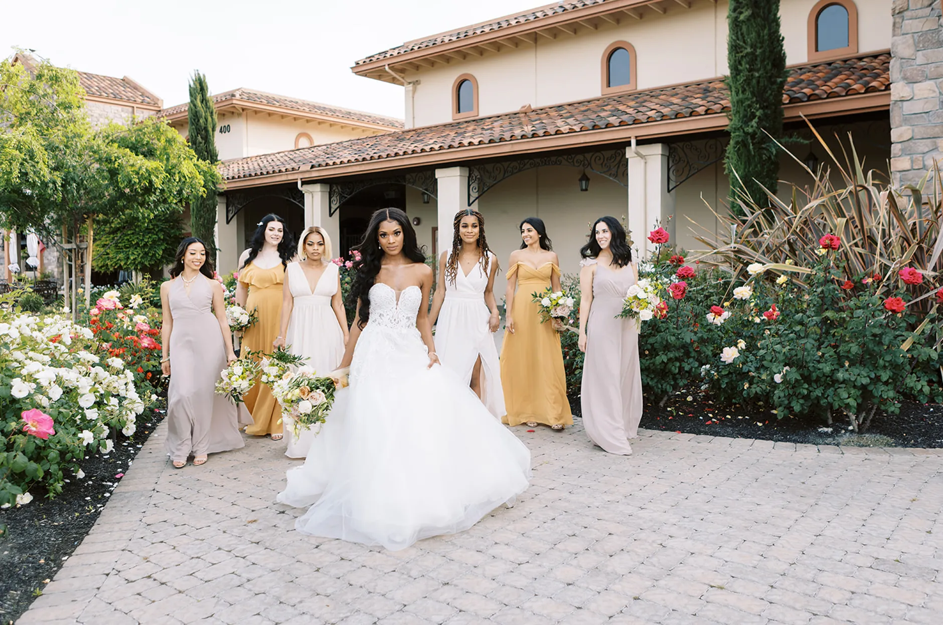 Bride and bridesmaids in gold and mauve dresses walking along a brick pathway past the venue rose garden