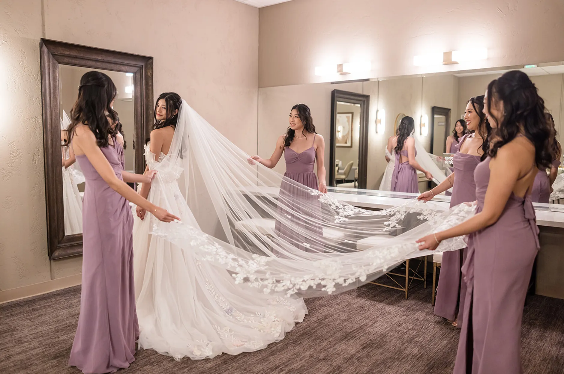 Bridesmaids in lavender dresses helping the bride spread her lace-trimmed cathedral veil in the bridal suite