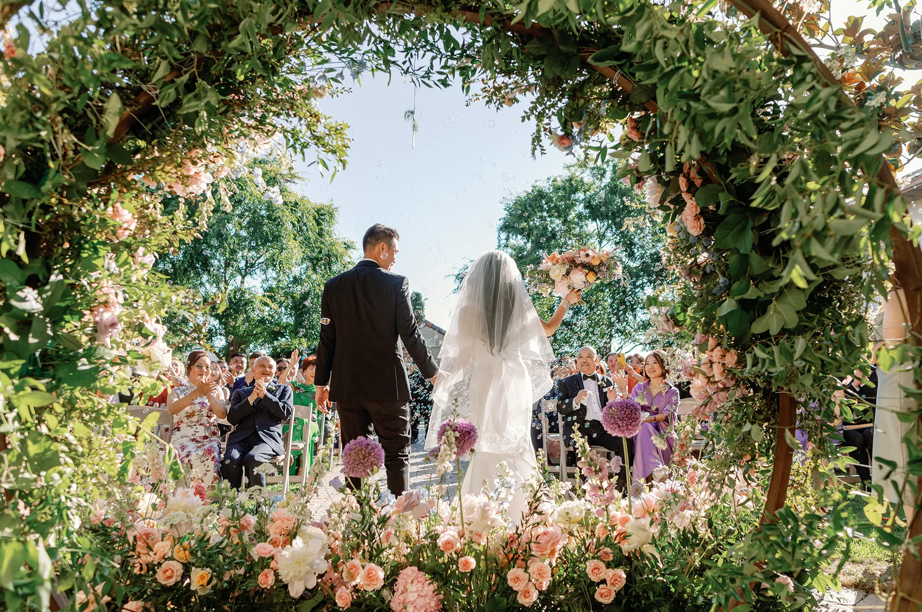 Couple walking hand in hand through a circular greenery arch lined with pastel flowers