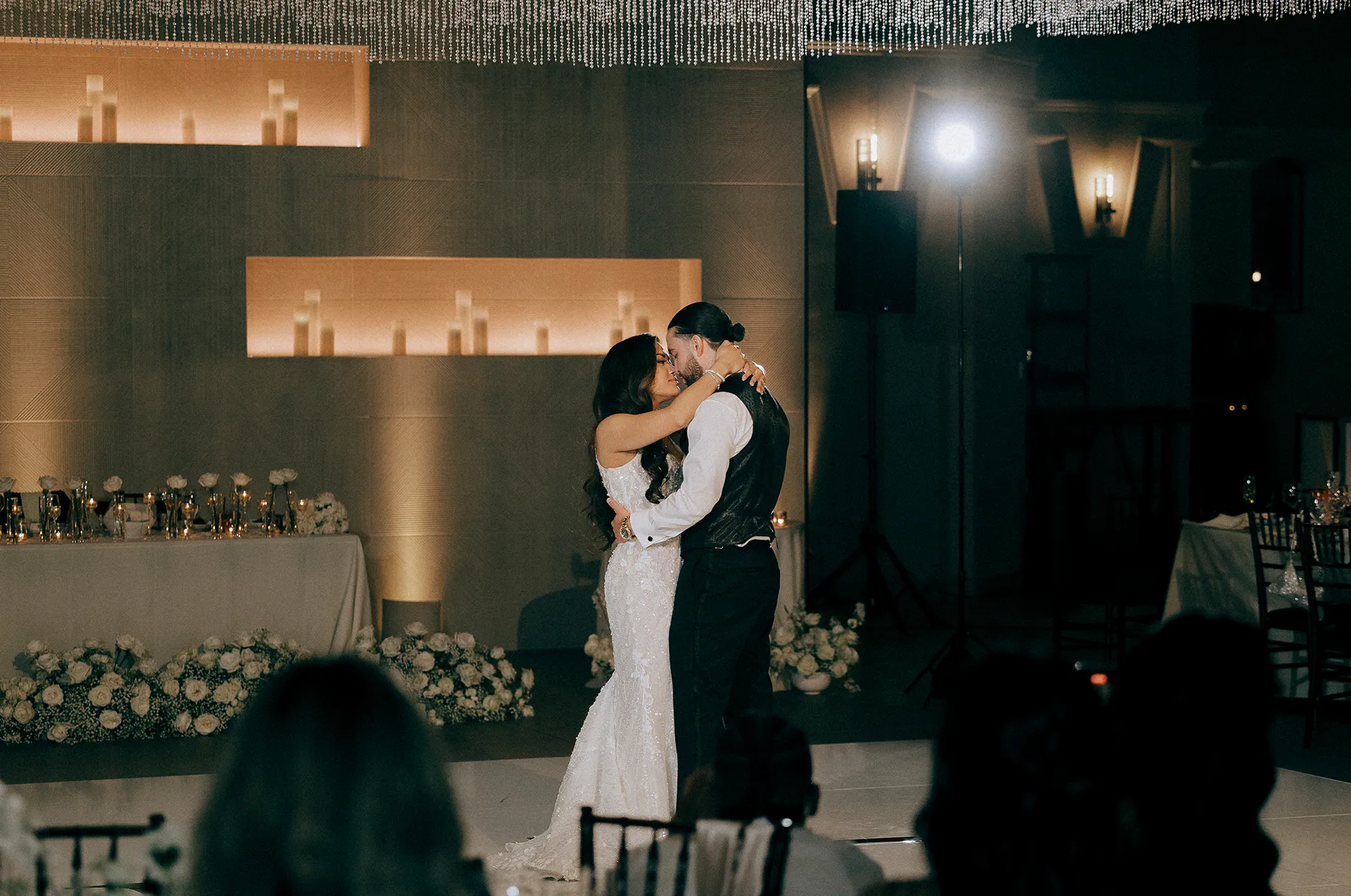 Couple sharing a kiss during their first dance under a canopy of string lights with white floral arrangements and candles on stage