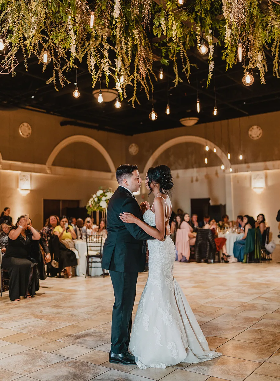 Couple sharing their first dance beneath a hanging greenery installation with Edison bulbs as guests look on