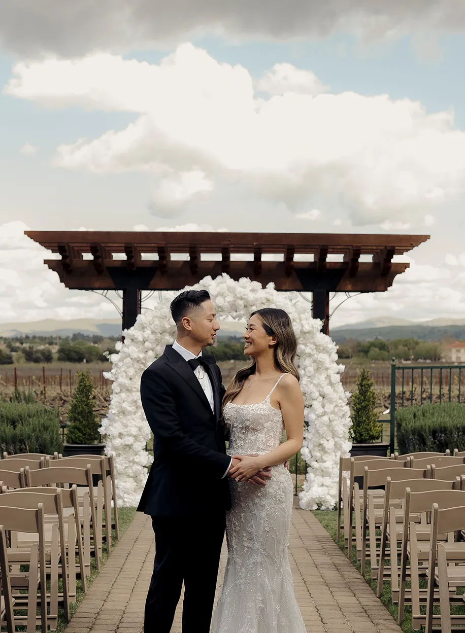 Couple gazing at each other beneath a wooden pergola and white balloon arch on the outdoor ceremony terrace with vineyard hills beyond