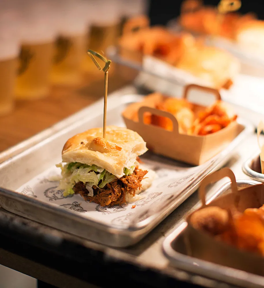 Pulled pork slider with lettuce on a metal tray alongside trays of chips and additional sliders