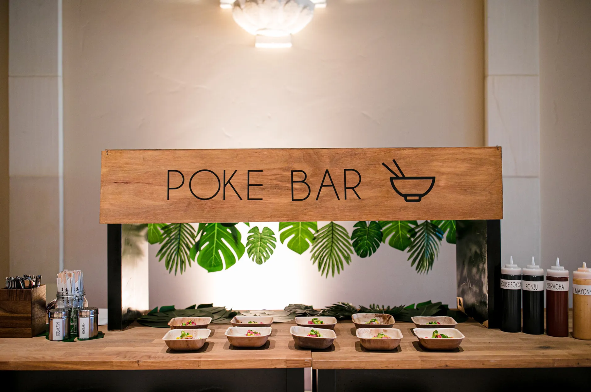 Poke bar station with a wooden sign, tropical leaf décor, and individual poke bowls lined up for guests