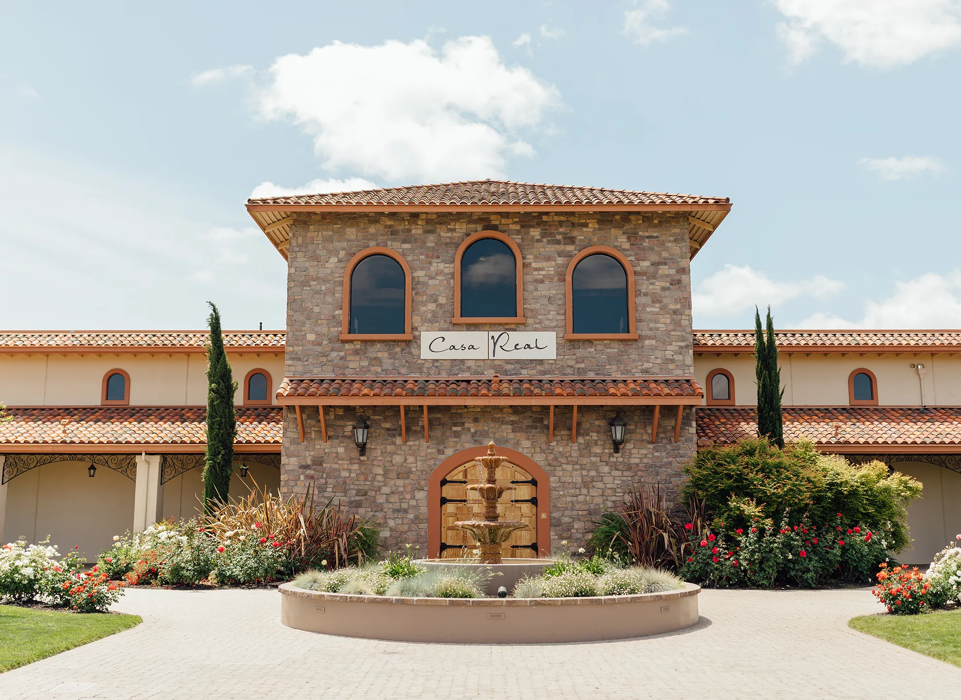 Stone facade of Casa Real with a terra-cotta roof, arched wooden doors, fountain, and Italian cypress trees