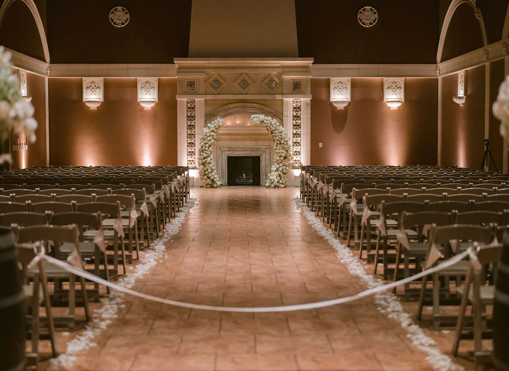 Amber Room set for a ceremony with rows of chairs, a petal-lined aisle, and a white floral arch by the fireplace