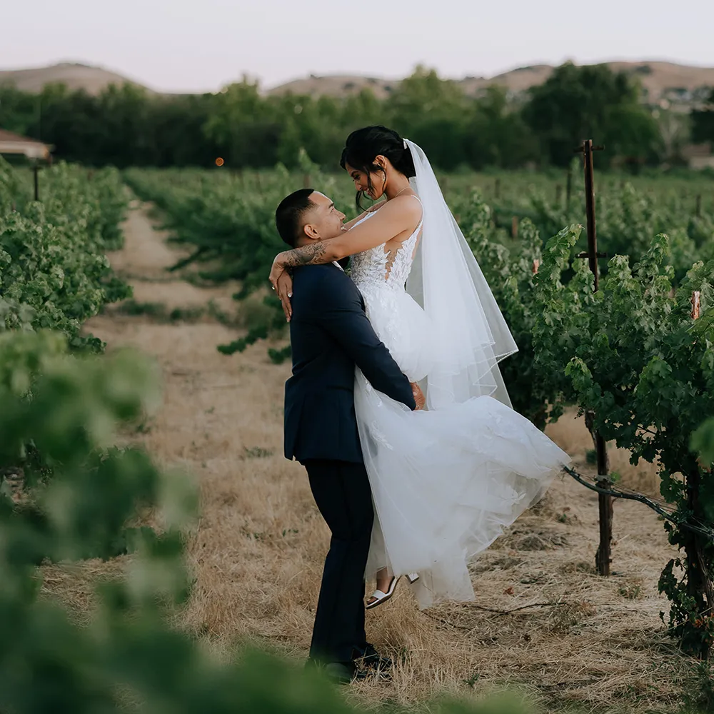 Groom lifting the bride in a vineyard row with rolling hills at sunset