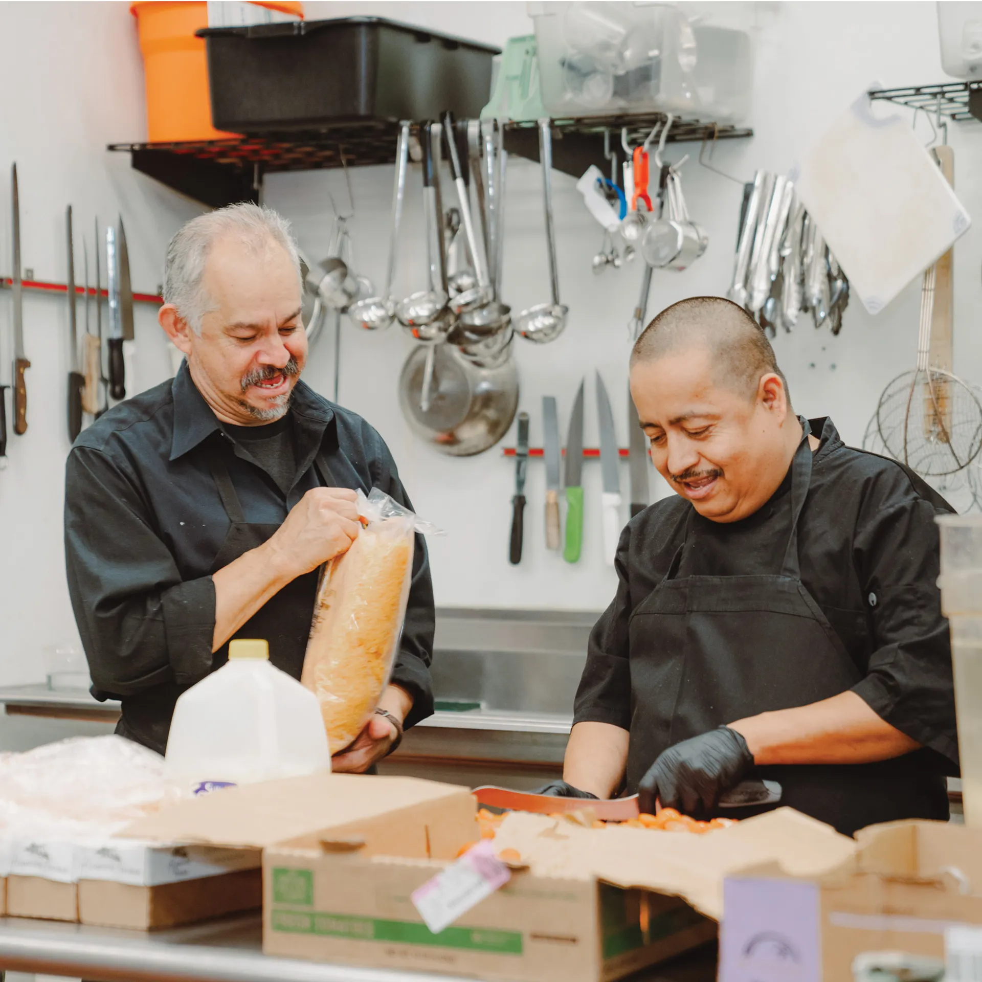 Two chefs in black uniforms smiling while prepping bread and ingredients in the kitchen