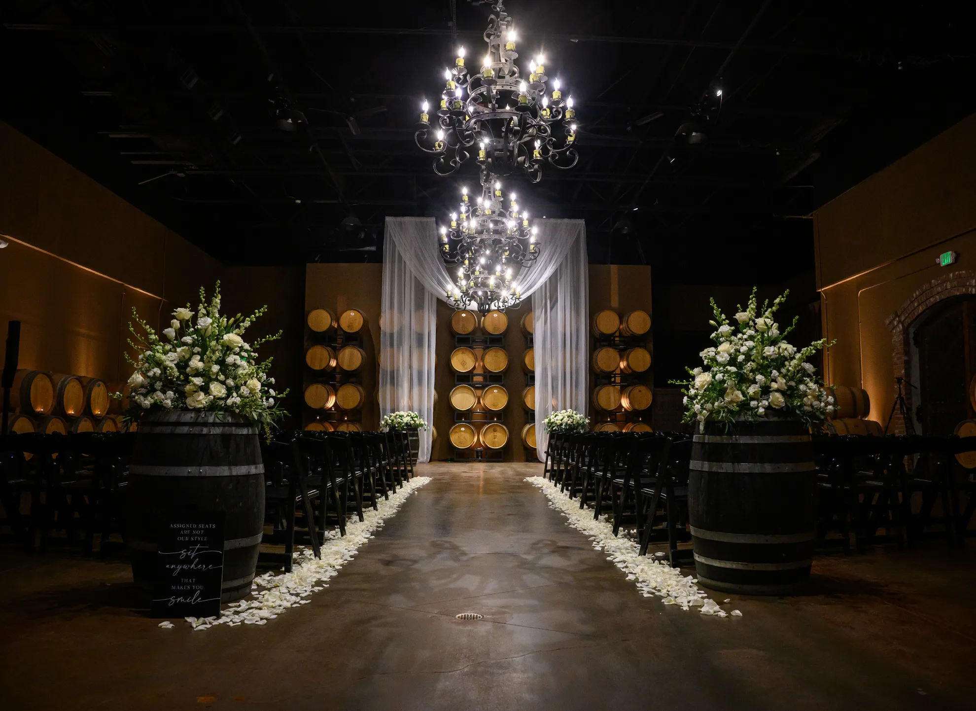 Estate Room ceremony with a petal-strewn aisle, white floral arrangements on wine barrels, and a wrought-iron chandelier