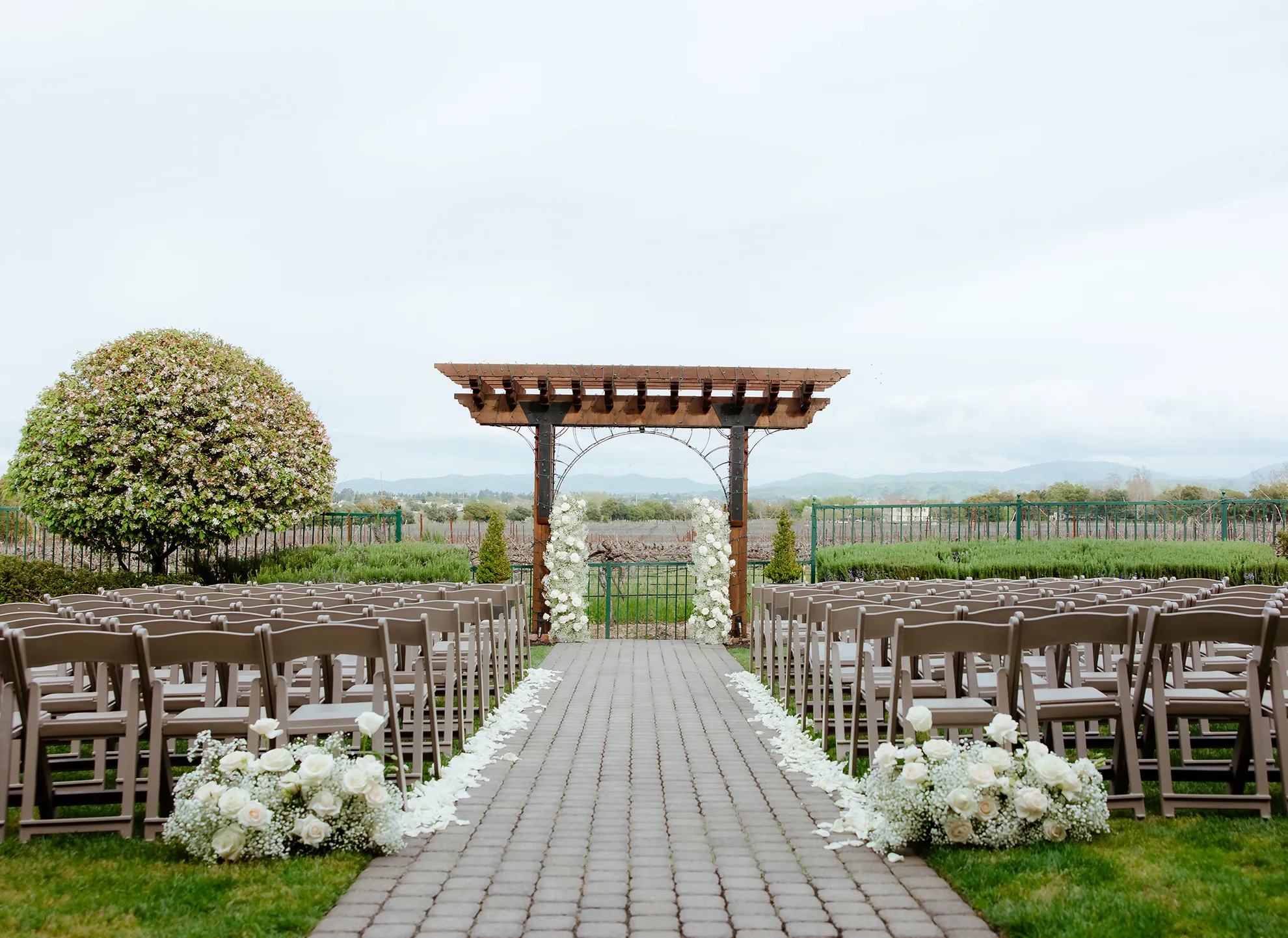Vineyard Lawn ceremony setup with rows of folding chairs, a white floral arch, and a wooden pergola overlooking the hills